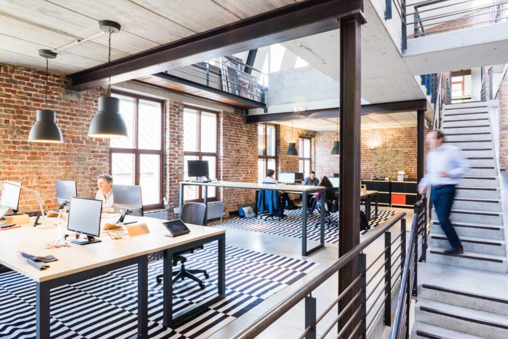 Modern office with brick walls, large windows, multiple desks with computers, and a person on a staircase.