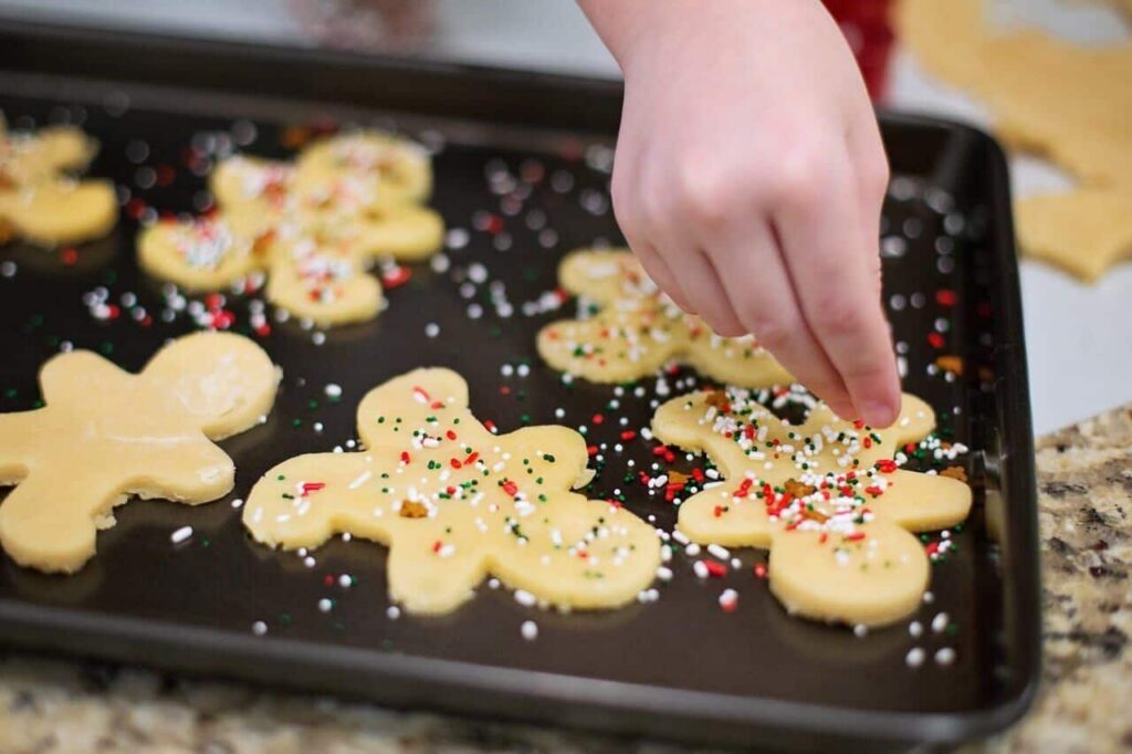 A hand sprinkles colorful red, white, and green sprinkles onto raw gingerbread-shaped cookie dough on a baking sheet.