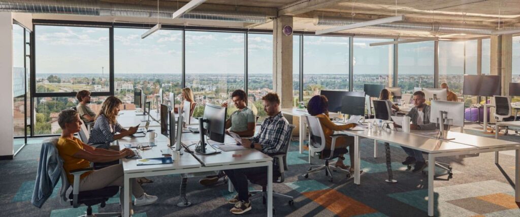 Modern open-plan office with people working at desks on computers. Large windows provide natural light and a city view, while a workplace management platform helps foster a bright, spacious, and collaborative atmosphere.