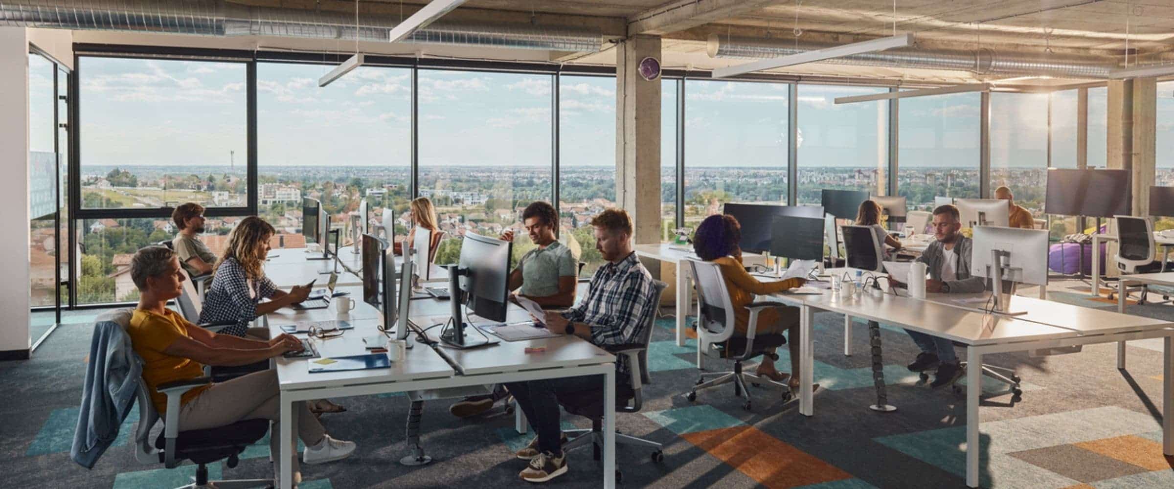 Modern open-plan office with people working at desks on computers. Large windows provide natural light and a city view, while a workplace management platform helps foster a bright, spacious, and collaborative atmosphere.