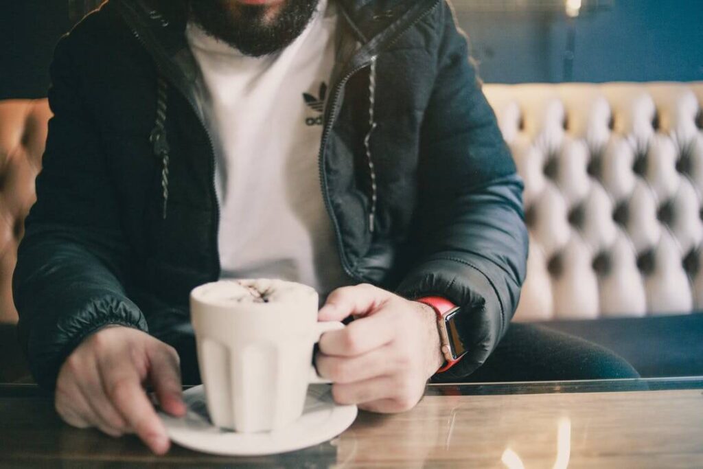 Person holding a cup of coffee in a cozy café setting, wearing a jacket and a smartwatch.