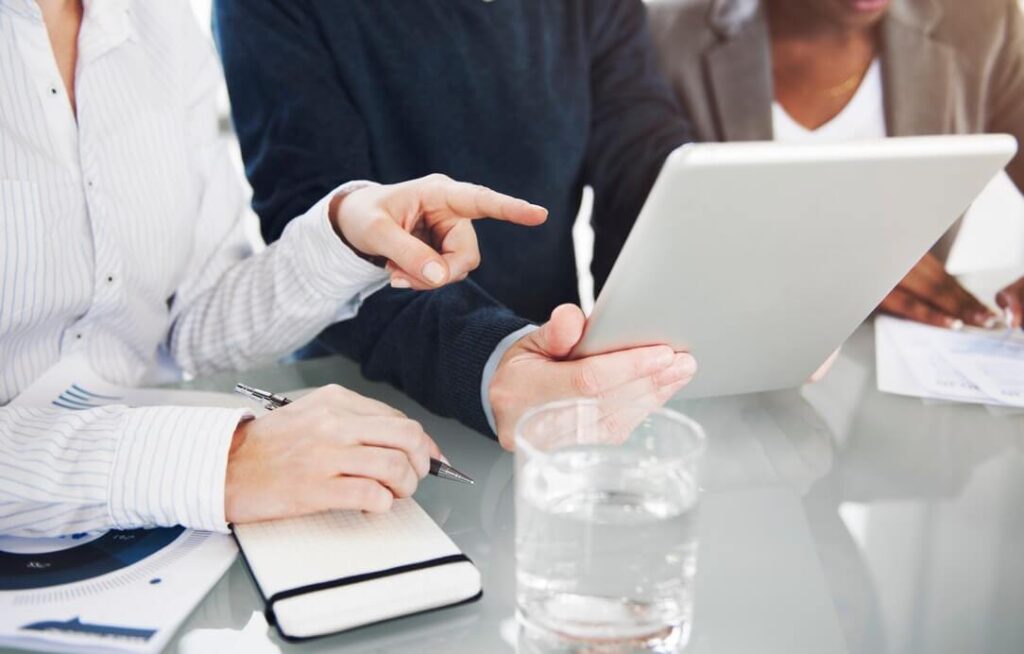 People sitting at a table, pointing at a tablet. Notepad, pen, and a glass of water are on the table.