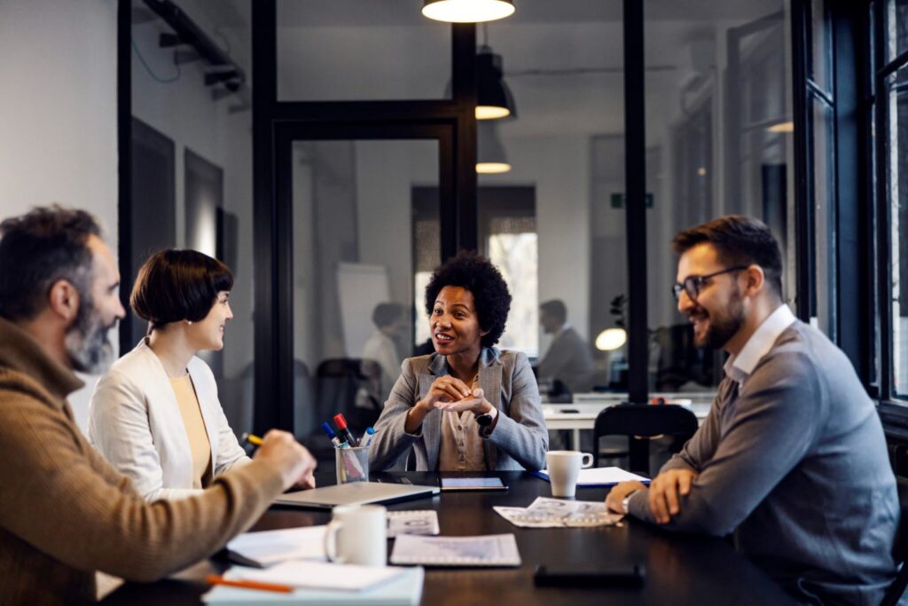 Four people in a meeting room, sitting at a table with papers and tablets, engaged in conversation and smiling.