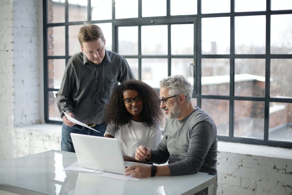 Three people from different generations in the workplace collaborate at a table, working together on a laptop in a bright, modern office with large windows. One person is sitting and typing, another is seated beside them, and a third stands, holding papers and looking at the screen.