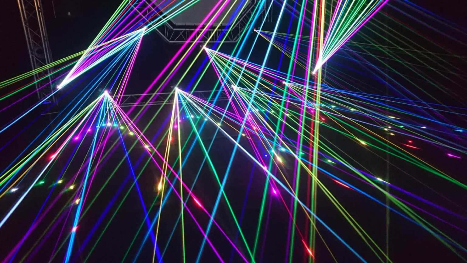 Multicolored laser beams crisscross in a dark space, creating sharp geometric patterns and a vibrant, energetic atmosphere.