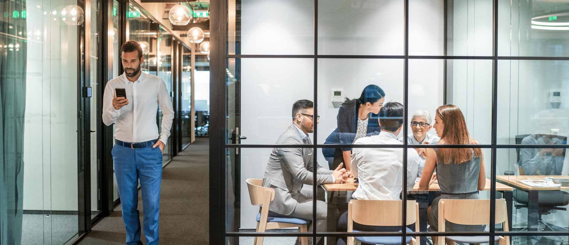 space utilization analysis. Colleagues sit in a glass-walled meeting room, a man walks past them in the hallway