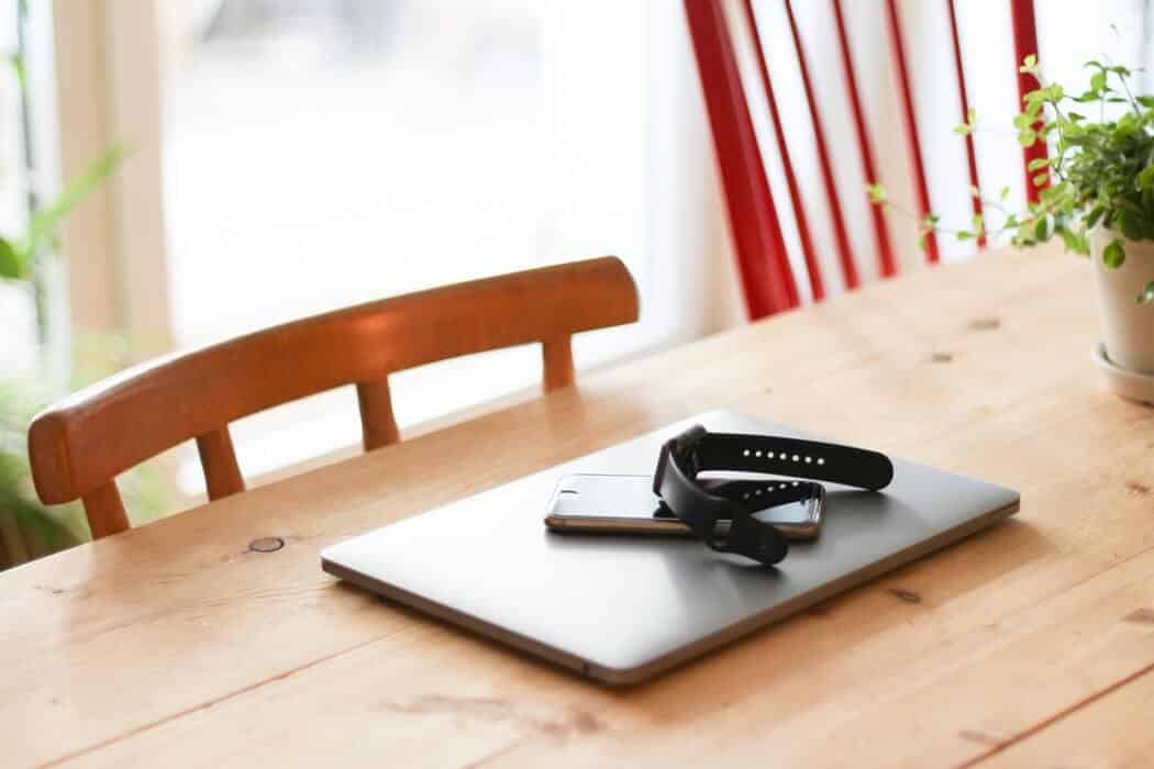 Smartwatch and smartphone on a closed laptop on a wooden table near a chair and a small potted plant.