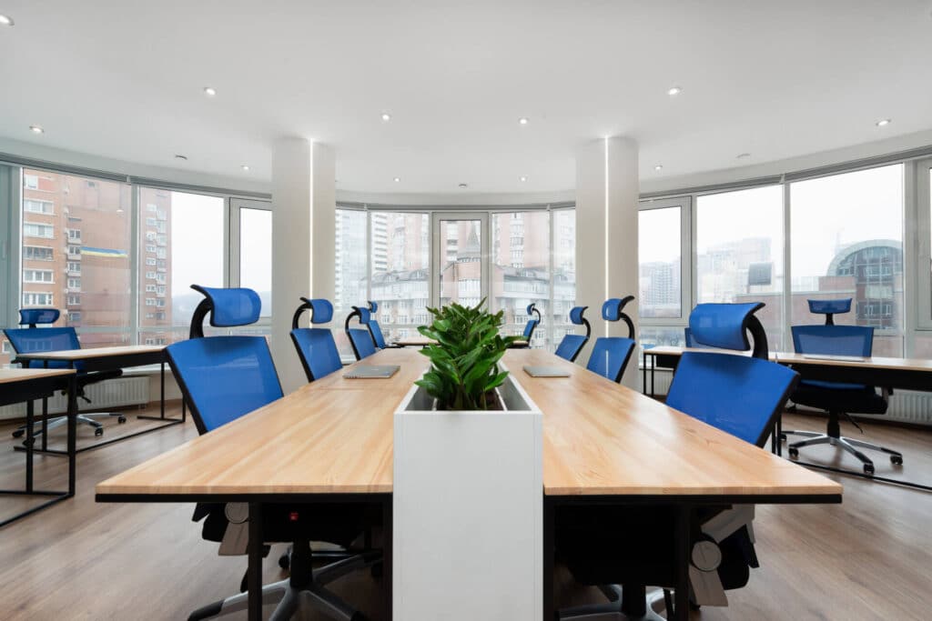 Modern office space with wooden desks, blue chairs, and a row of potted plants, set against large windows.
