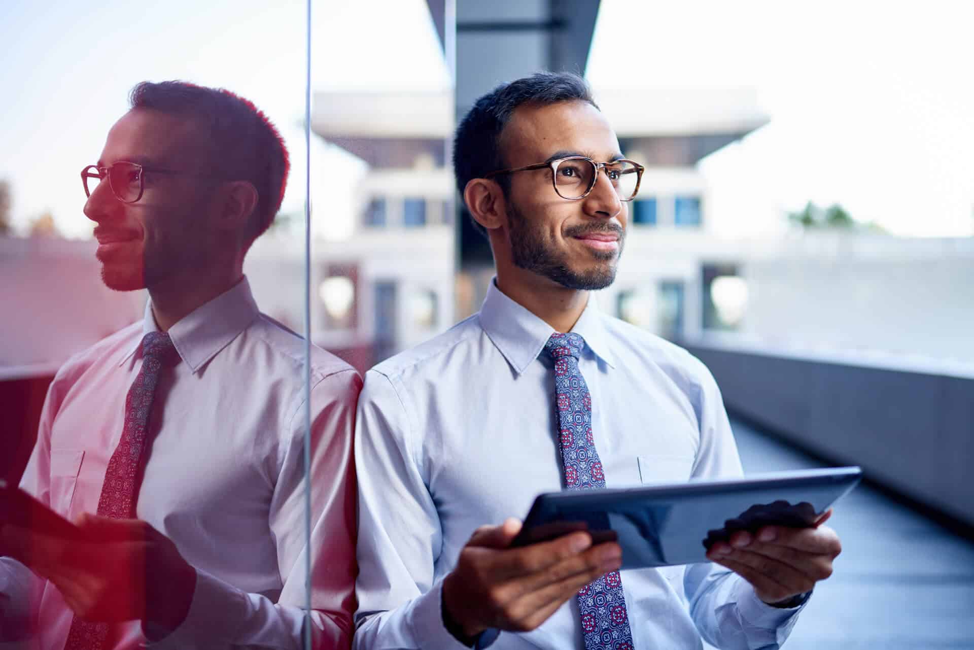 Man in a tie holding a tablet, smiling, stands next to a reflective glass wall with a modern building in the background.