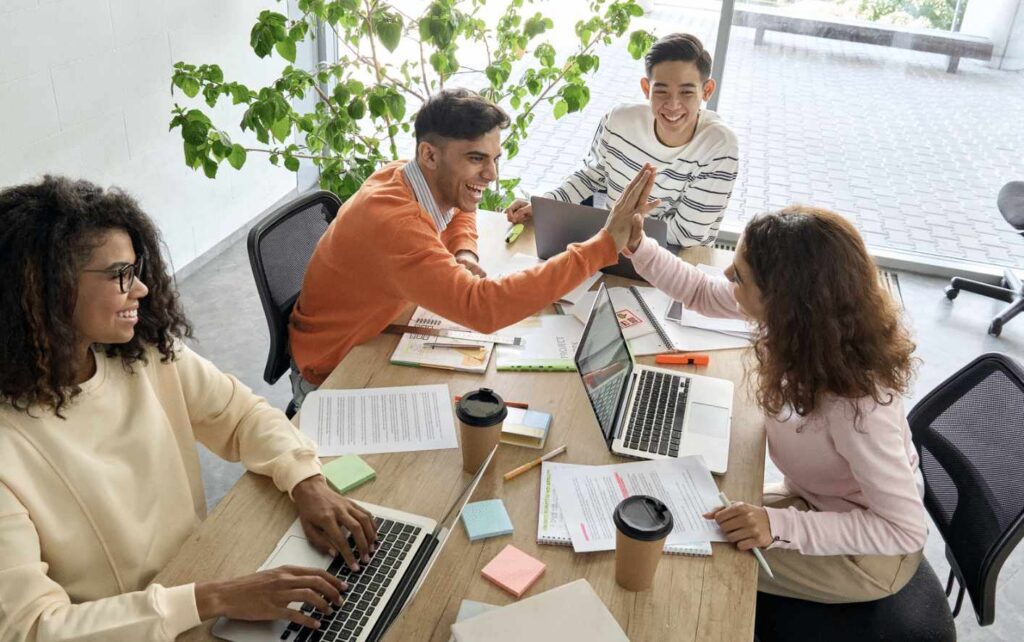 Four young adults sit at a table with laptops and papers, embracing the excitement of the return to office. Two of them high-five while the others smile, enjoying their time together. Coffee cups and sticky notes are scattered across the table, and a large plant is in the background.