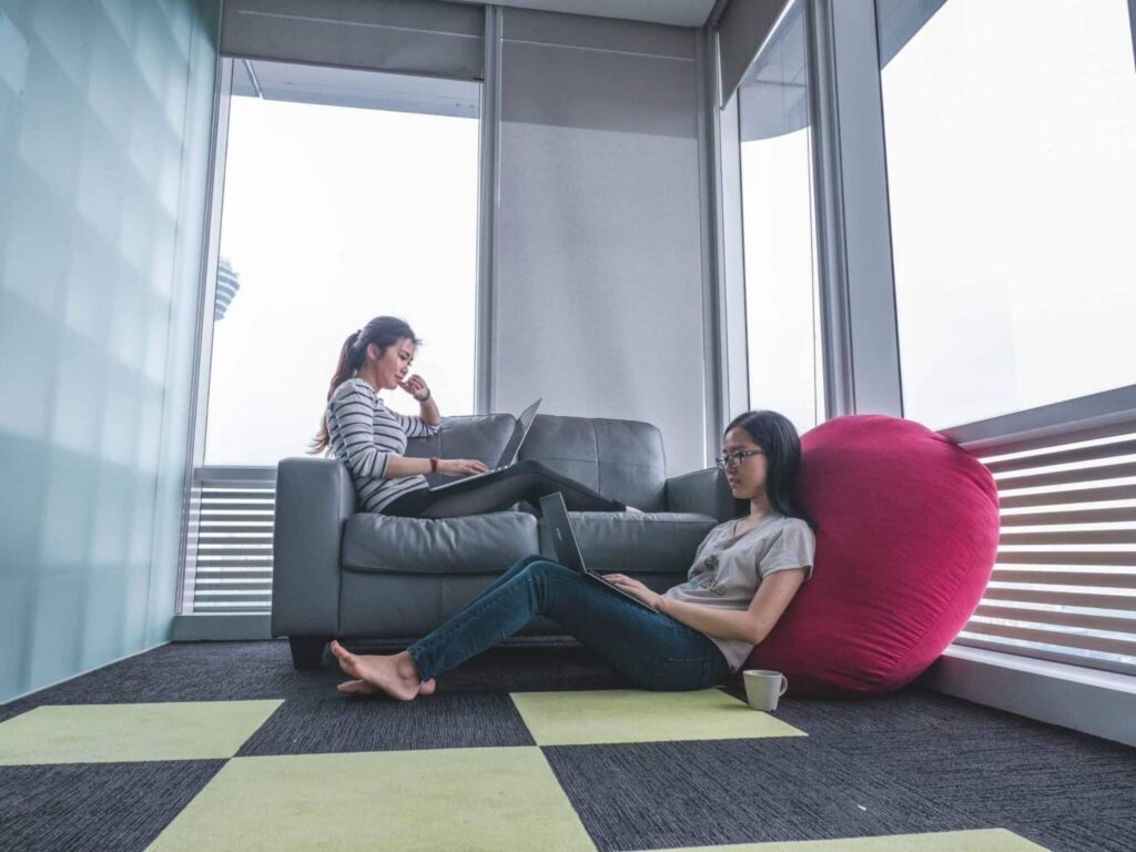Two women using laptops, one sitting on a couch and the other on the floor leaning against a pink beanbag, demonstrate agile work examples in a modern office with large windows and patterned carpet. A coffee cup is on the floor nearby.