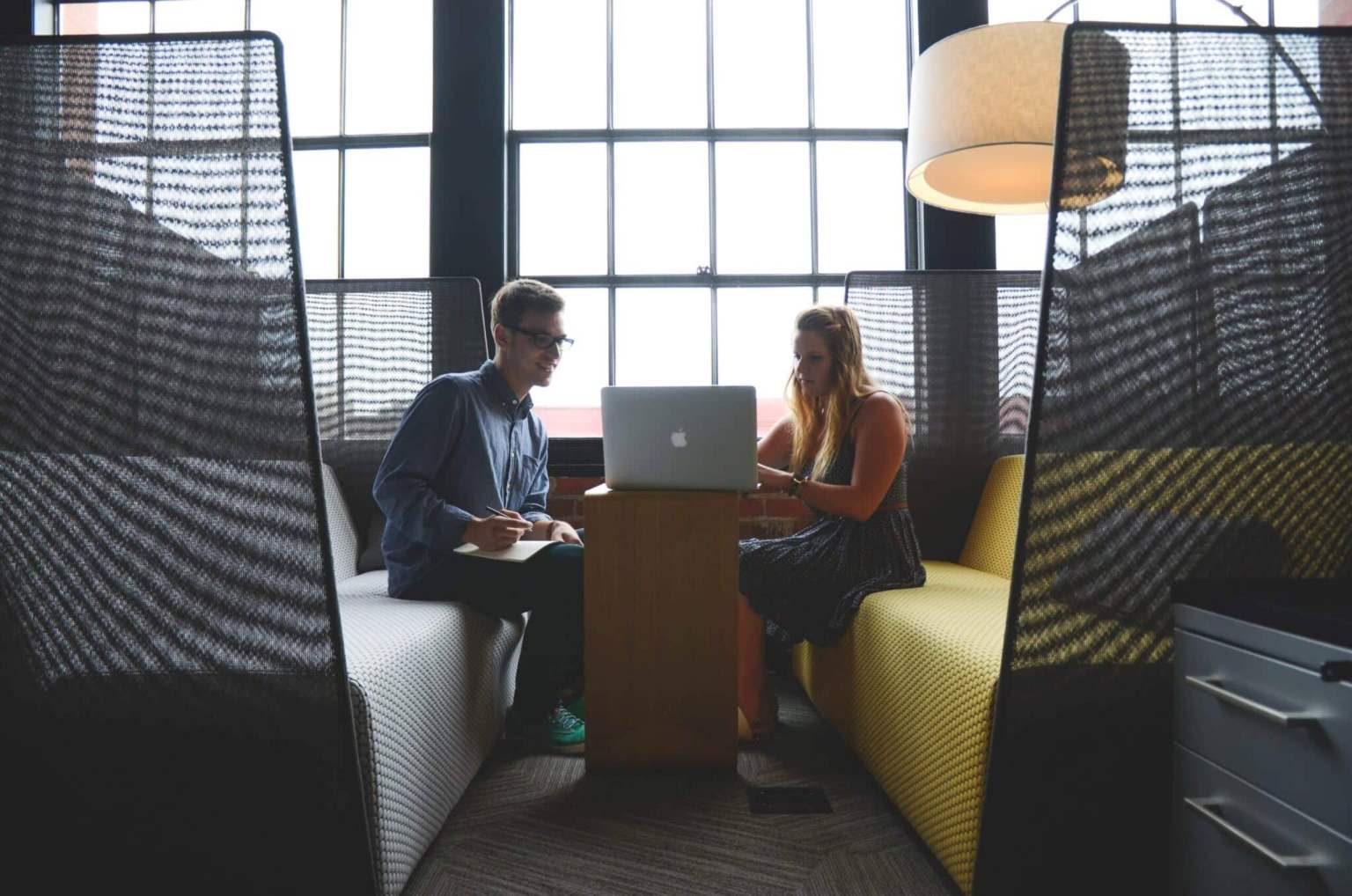 Two people sit across from each other in a modern office booth, working together at a laptop on a small wooden table, with large windows and soft lighting in the background—a collaborative scene highlighting the importance of effective office facility management.