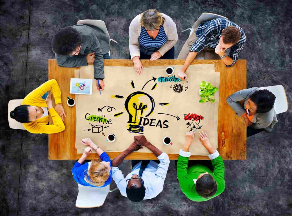 A diverse group of people sit around a table viewed from above, brainstorming with a large paper showing a lightbulb and the words Ideas, Creative, Think, and Design—perfect inspiration for unique conference room names. Coffee cups and plants are scattered on the table, adding to the creative atmosphere as they discuss potential room names that spark innovation.