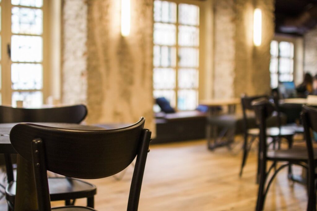 Cozy cafe interior with wooden chairs and tables, soft lighting, and large windows letting in natural light.