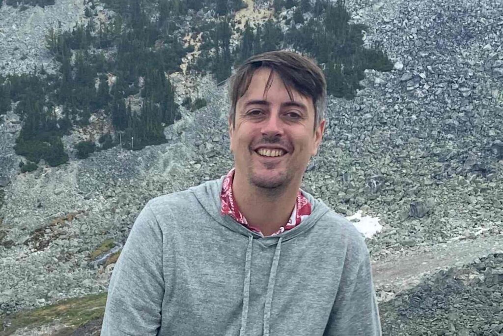 Smiling person in a gray hoodie standing in front of a mountainous landscape with scattered trees and rocks.
