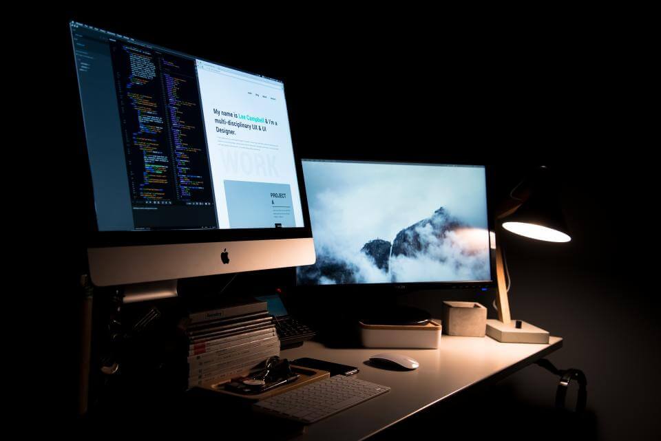 Dimly lit desk with two monitors displaying code and a mountain scene, a lamp, and books in a dark room.