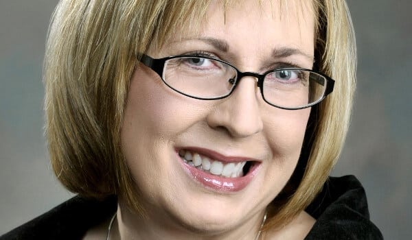 Smiling person with short blonde hair, glasses, and a dark top against a neutral background.