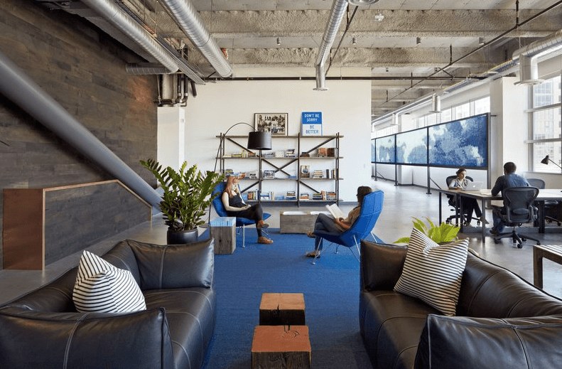 Modern open office space with two people talking in blue chairs on a blue rug, others working at desks, bookshelves and large windows in the background, and black sofas with striped pillows in the foreground.