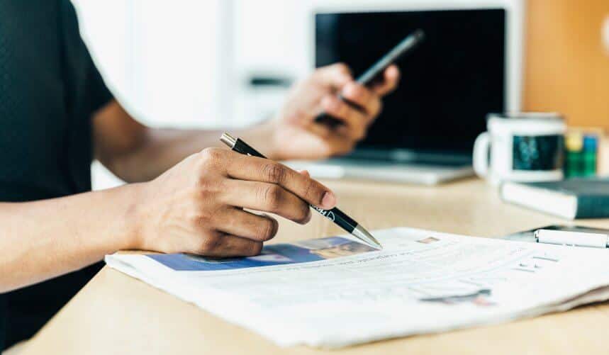 Person reading a newspaper, holding a pen and a smartphone, with a laptop and coffee mug nearby.