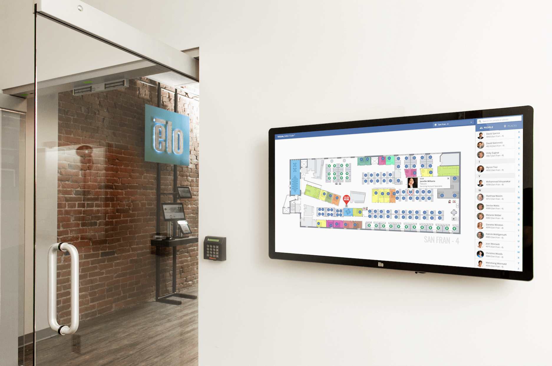 A digital office floor map is displayed on a wall-mounted screen next to a glass door with a handle. A keypad and an elo sign are visible through the door in an exposed brick hallway.
