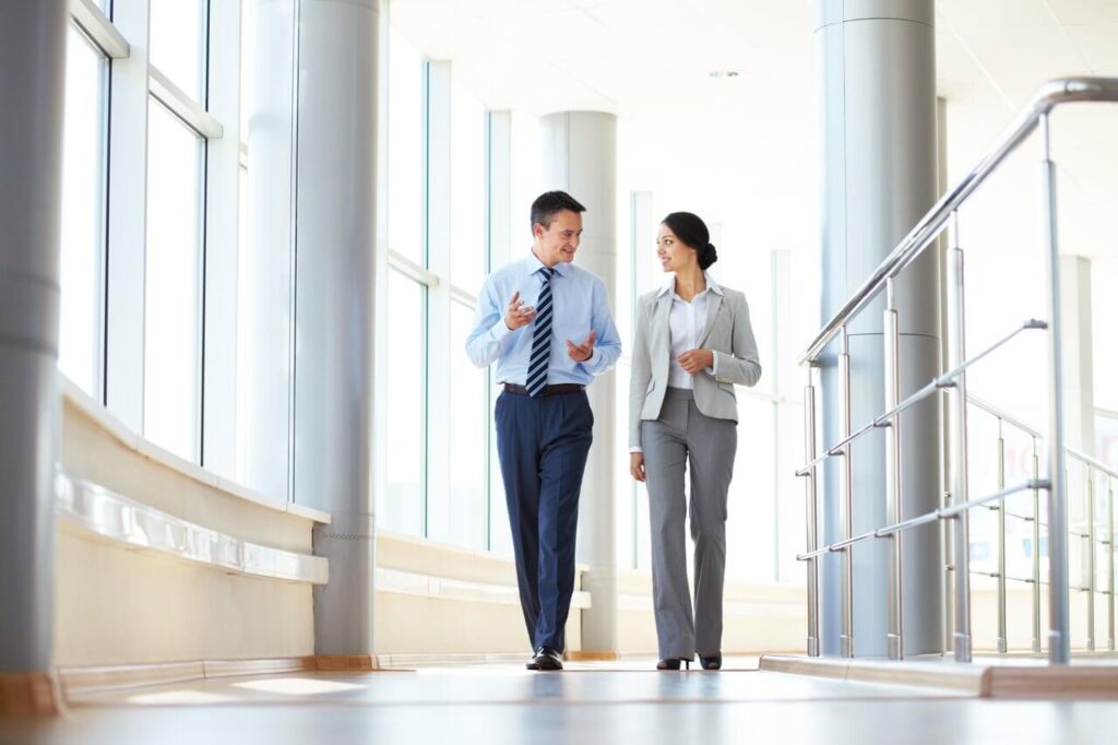 Two professionals in suits walking and talking in a bright hallway with large windows and metal railings.