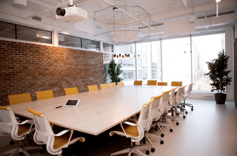 A modern conference room designed as an inspiring office space, featuring a large wooden table, white and yellow chairs, brick wall, hanging lights, a tablet on the table, potted plants, and large windows letting in natural light.