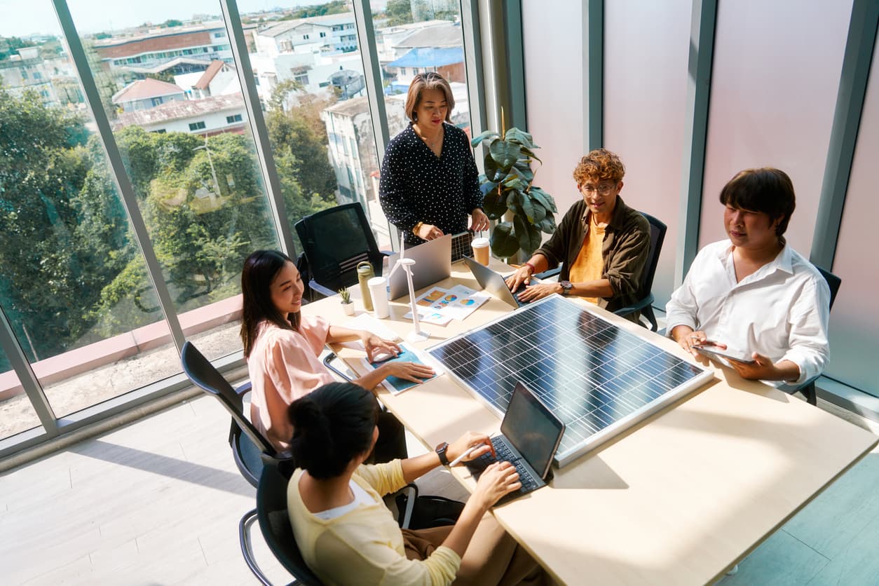 A diverse team attentively discusses renewable energy, with a large solar panel on the table, in a modern office.