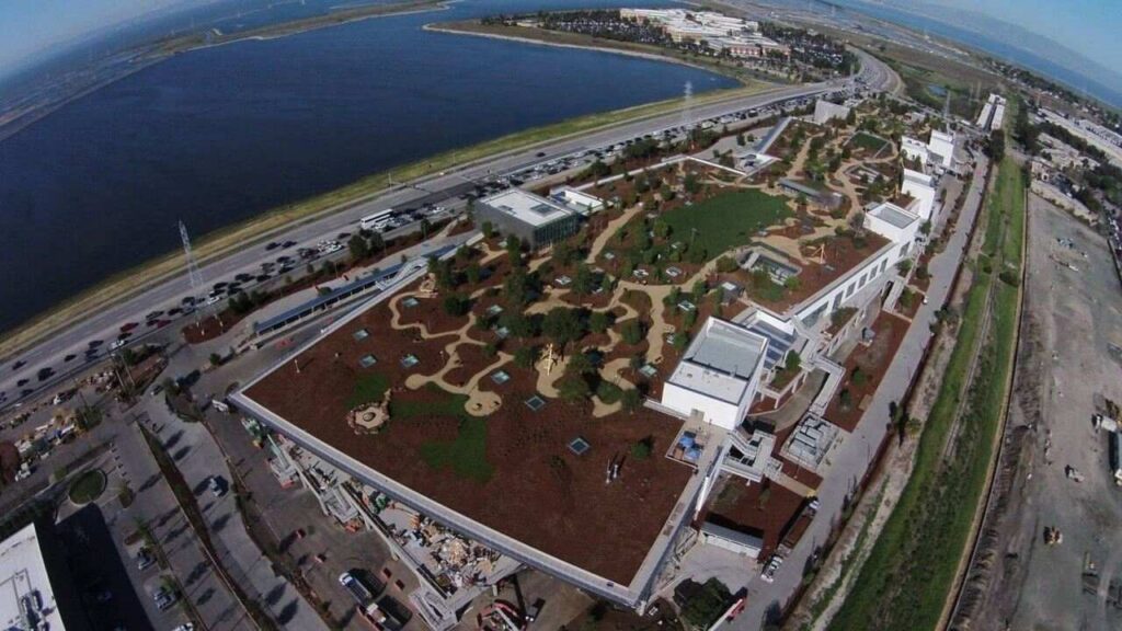 Aerial view of a large building with a rooftop garden featuring trees, pathways, and green spaces, located near a body of water and surrounded by roads and industrial areas.