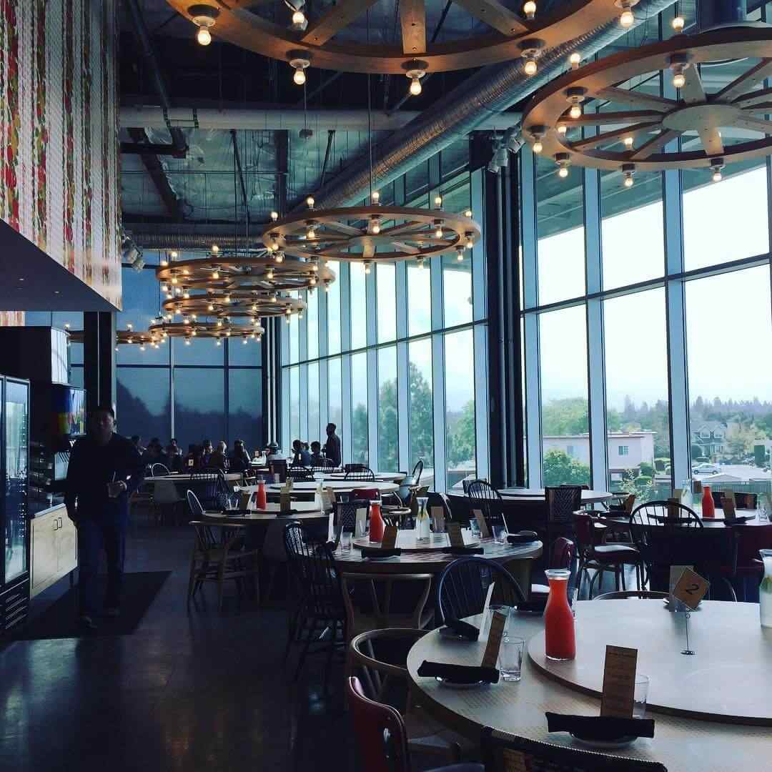 Modern restaurant with large floor-to-ceiling windows, round tables set with condiments, and unique circular ceiling lights. Several people are seated inside, with an outside view of trees and buildings.