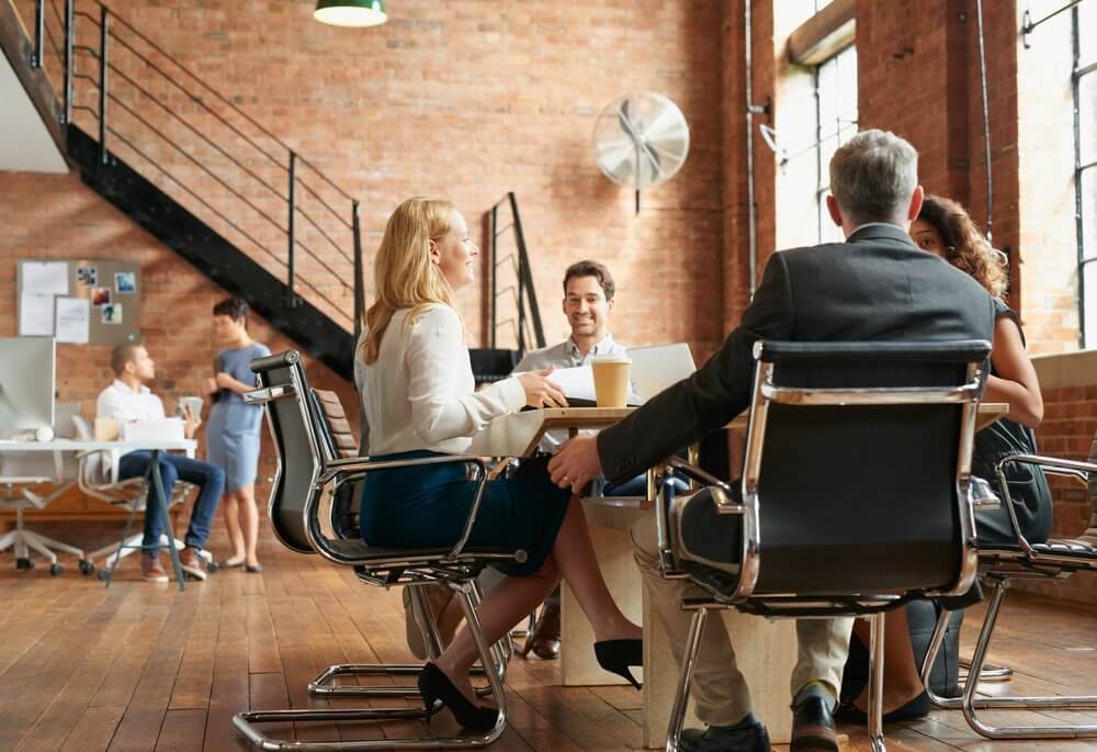 People sitting at a table in a modern office with brick walls and large windows, engaged in conversation.