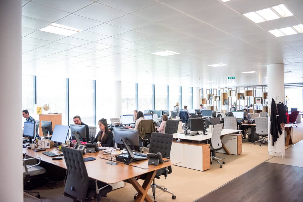 Open-plan office with people working at desks, computers, and large windows providing natural light.