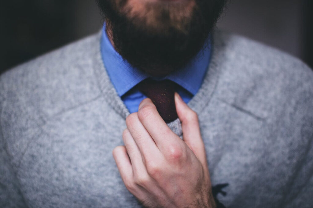 A person adjusts a dark tie, wearing a blue shirt and gray sweater.