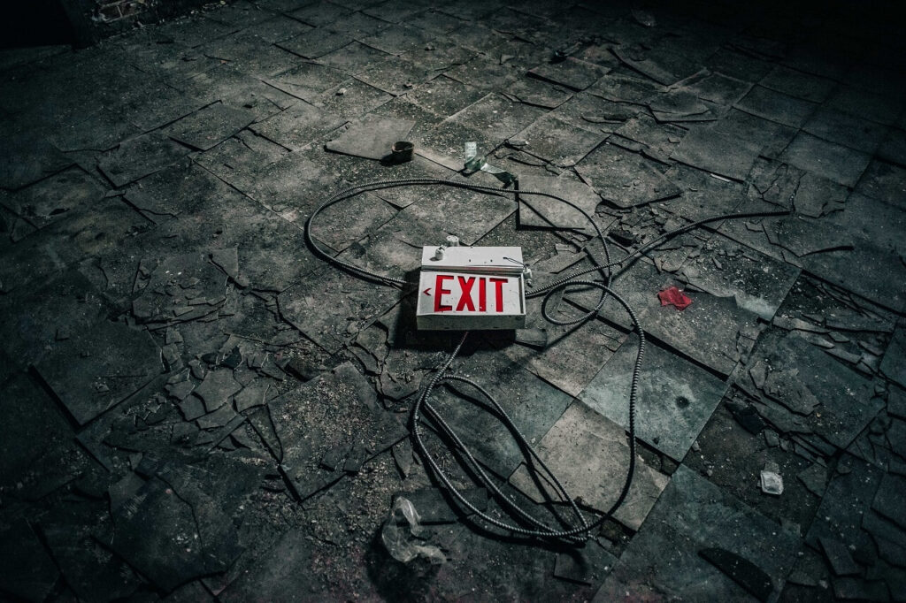 A broken "EXIT" sign lies on a cracked, dirty floor with scattered debris and wires.