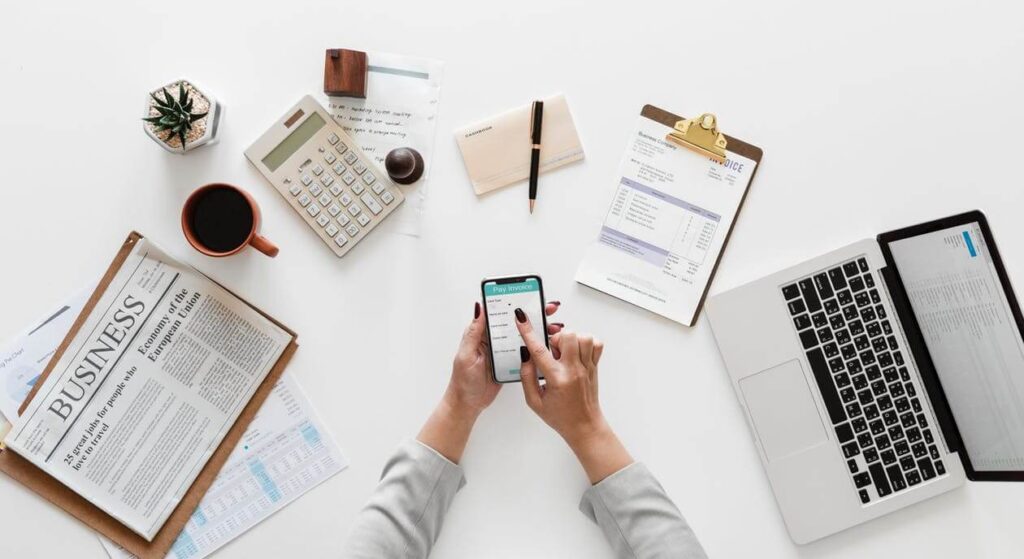 Person using a smartphone at a desk with a laptop, newspaper, coffee, calculator, and documents.