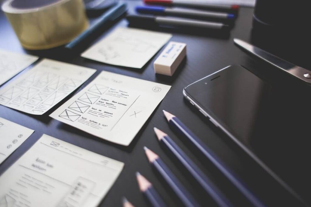 Desk with design sketches, pencils, smartphone, eraser, and pens on it.