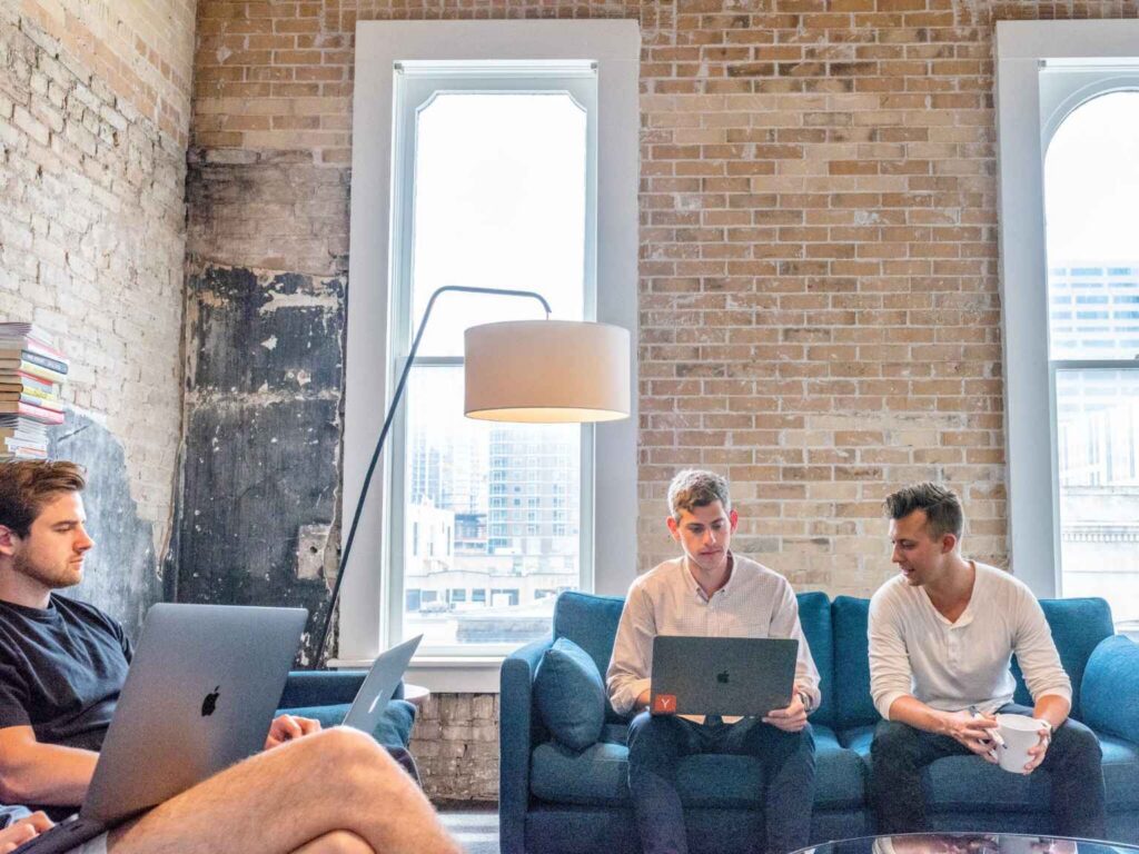 Four people from different generations in the workplace sit in a modern office lounge with laptops, discussing work. Exposed brick walls, tall windows, and a floor lamp create a relaxed, collaborative atmosphere.