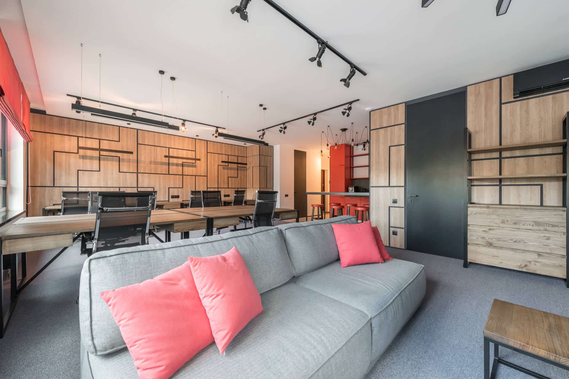 Modern office with a gray sofa, pink cushions, wooden desks, black chairs, and unique geometric wall design.