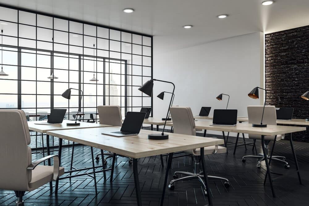 A modern office with wooden desks, laptops, and lamps, featuring large windows and black metal framing.