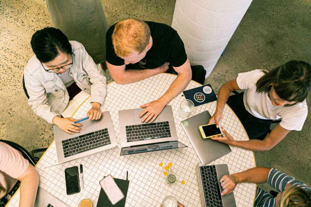 Four people work together at a round table with laptops, notebooks, a phone, and drinks, seen from above. They appear focused and engaged in a collaborative task—an ideal scene for office neighborhood examples illustrating how teams can connect using neighborhood software to foster effective collaboration.