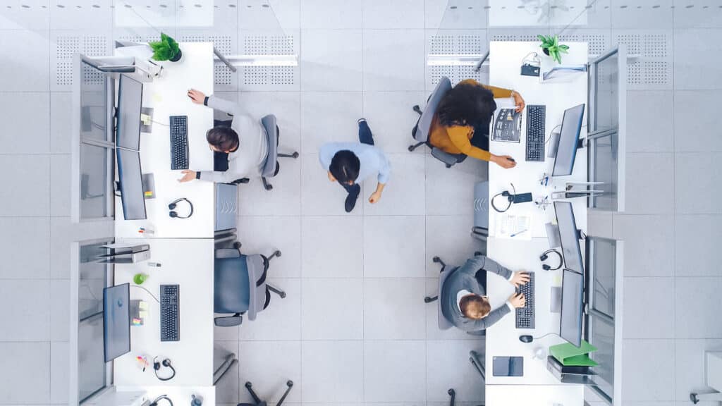 Top view of an office with people working at desks, computers, and a person walking in the aisle.