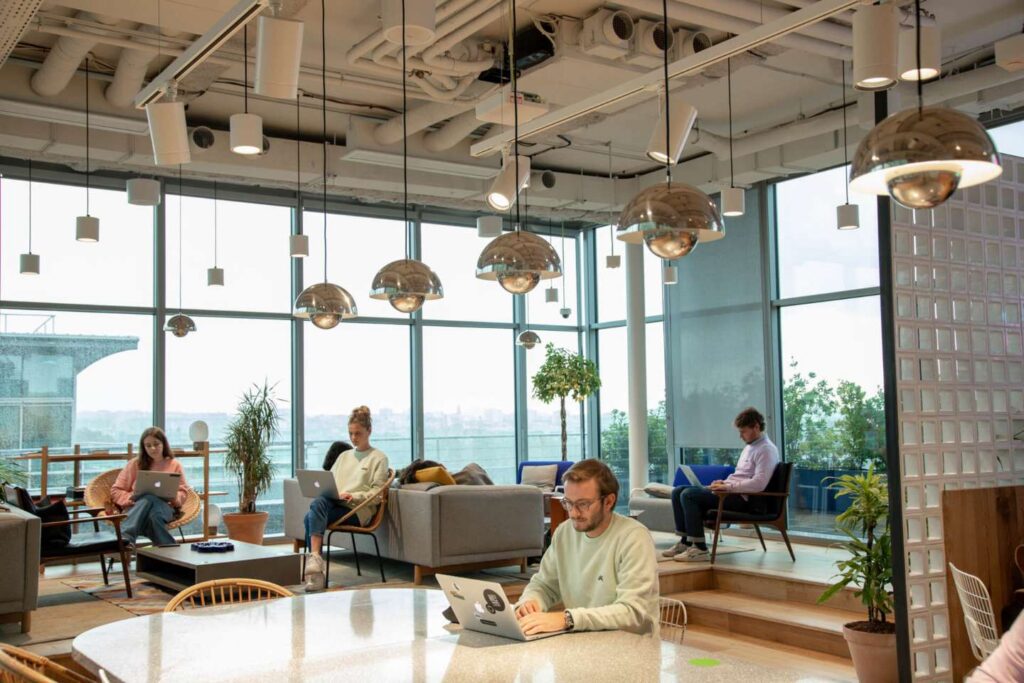 Four people work on laptops in a modern, open office with large windows, hanging lights, and potted plants. Embracing a free address workplace concept, each person chooses their own spot to work for the day. Outside, a city skyline is visible through the glass. The space feels bright and collaborative.