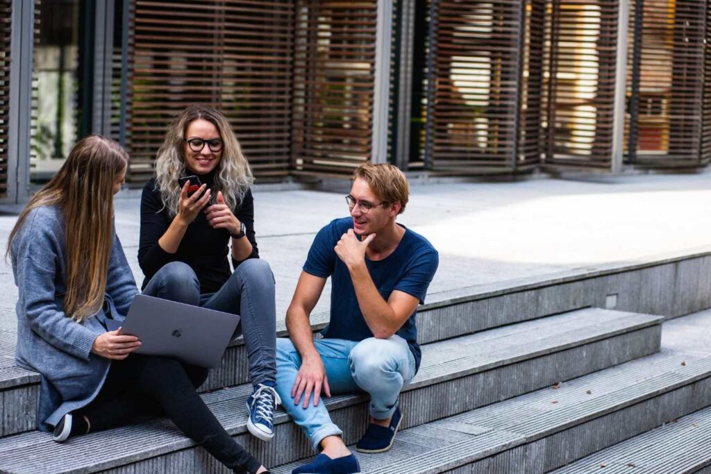 Three young adults sit on outdoor steps, smiling and chatting. One holds a laptop, another shows something on a phone—perhaps sharing ideas about workplace IoT—and the third looks on, engaged. They appear relaxed in a modern urban setting.