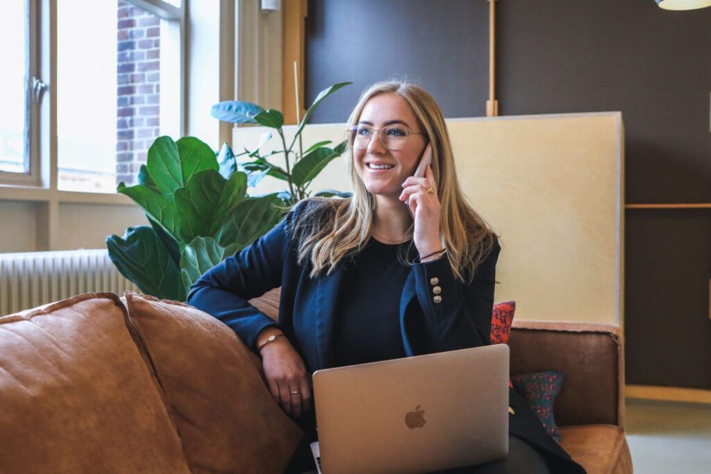 A woman with glasses sits on a couch, smiling while talking on the phone. She has a laptop on her lap and is in a modern, well-lit room with large windows and a green plant nearby, reflecting the dynamic environment shaped by multiple generations in the workplace.
