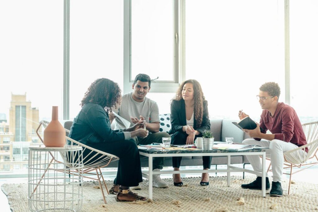 Four people sit around a small table in a bright room, discussing and writing in notebooks.