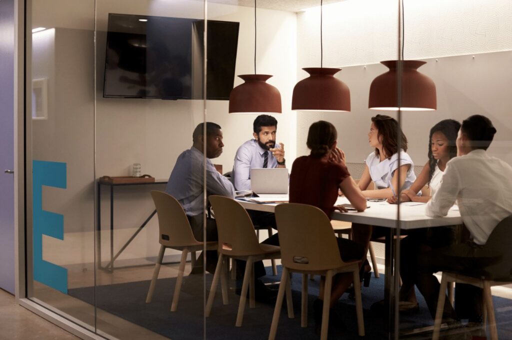 Six people having a meeting around a table in a modern office, seen through a glass wall.