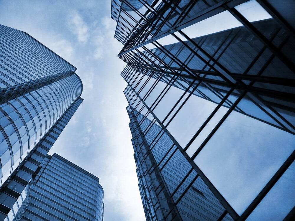 Workplace Unplugged, Change Management with David Slight of Quora Consulting Low-angle view of modern glass skyscrapers reflecting the sky, with blue-tinted windows and metal frames, creating a geometric and contemporary cityscape against a partly cloudy sky.