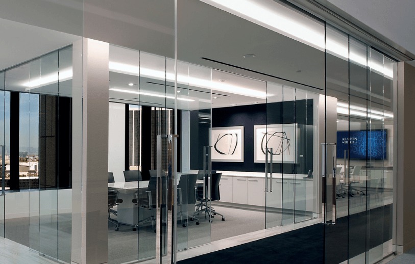 Modern office conference room with glass walls, a long table surrounded by chairs, abstract art on the walls, and a large monitor displaying blue graphics, with city buildings visible through the windows.