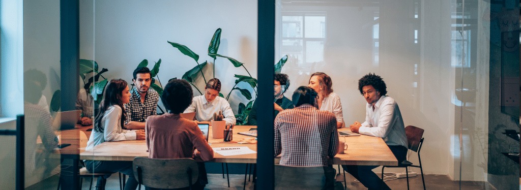A group of people sit around a large table in a modern office meeting room, discussing and working on laptops. Green plants decorate the room, and natural light comes in through glass walls. The seamless coordination of their gathering is thanks to an efficient conference room booking system that keeps meetings organized and hassle-free.