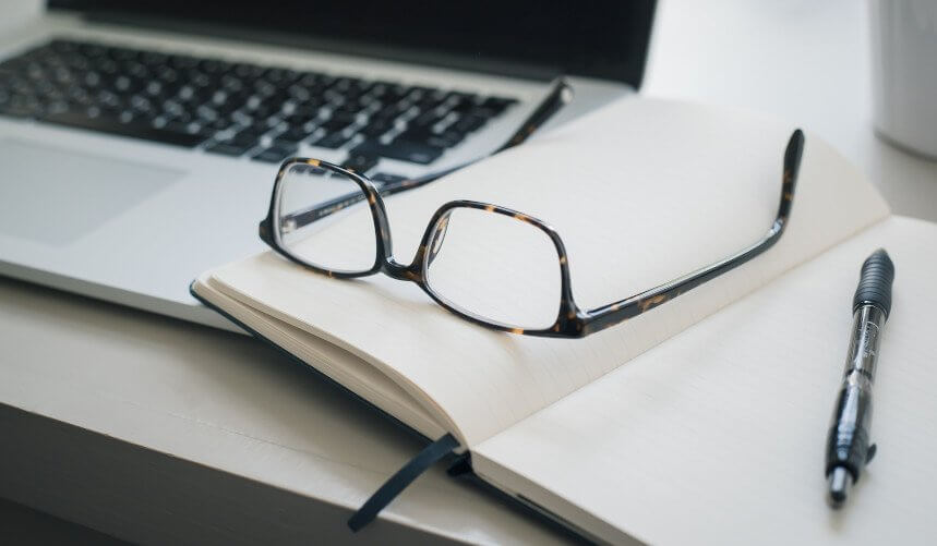 Open notebook with glasses and pen on top, next to a laptop on a desk.