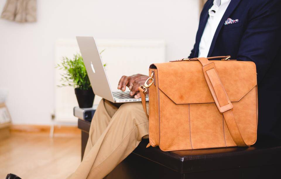 Person with laptop and tan leather bag, sitting on a bench in a modern setting.
